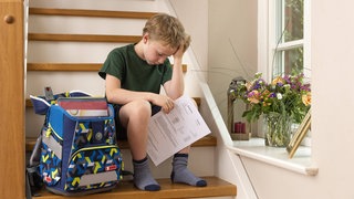 Ein Grundschüler sitzt traurig mit seinem Zeugnis auf der Treppe: Studien zeigen, dass vom Sitzenbleiben niemand profitiert. Dafür sind die Ehrenrunden oft demotivierend und obendrein ganz schön teuer. Einige Bundesländer sind schon ausgestiegen.