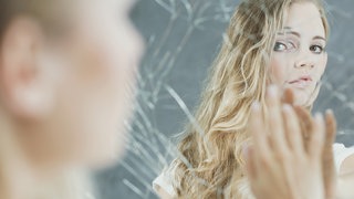 Junge Frau schaut in einen kaputten Spiegel: "Warum hat es mich erwischt?" Das fragen sich viele Kranke. Der Zusammenhang zwischen Psyche und Krankheit beschäftigt Forschende.