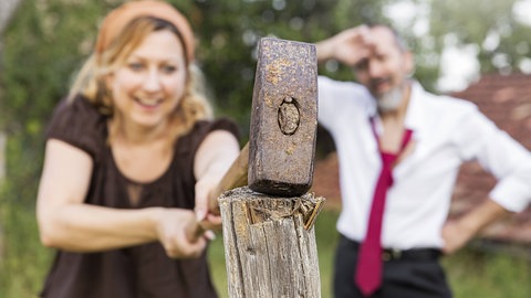 Eine Frau schlägt einen Zaunpfahl ein, ein Mann mit Krawatte streicht sich im Hintergrund erschöpft über die Stirn: Immer mehr Stadtmenschen zieht es raus aufs Land