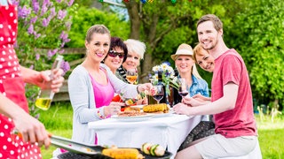 Grillfest im Garten: Sechs Menschen sitzen am Tisch, eine Person grillt: Einerseits werden Nachbarn immer zwischen Anonymität, Konflikt, Freundschaft und Gemeinschaft pendeln. Andererseits leben einige – auch mithilfe moderner Stadtplanung – inzwischen vor, was sie in guter Nachbarschaft gewinnen können.