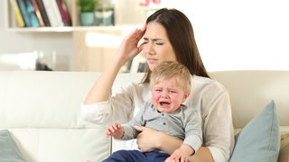 Gestresste Mutter hält weinendes Kleinkind auf dem Schoß: Perfekt organisiert und gelassen, vormittags erfolgreich im Beruf und nachmittags Spielkameradin und Hausaufgabenbetreuerin. Die Anforderungen an eine "gute Mutter" sind heute groß – und oft widersprüchlich. Das idealisierte Mutterbild prägt auch die hohen Ansprüche, die viele Mütter an sich selbst stellen. Der Mythos der angeborenen, bedingungslosen Mutterliebe sitzt tief. 
