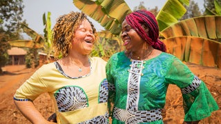Zwei junge afrikanische Frauen, eine Frau hat Albinismus, stehen im Freien und lachen sich an: Menschen mit Albinismus verfolgt in schwarzafrikanischen Ländern wie Sambia tiefsitzender Aberglaube; sie sind von Geburt an stigmatisiert. Die Diskriminierung wurzelt in altem Aberglauben und ist schwer auszurotten. Doch die betroffenen Menschen wehren sich gegen das Stigma.