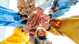 Junge Menschen stehen in einem Kreis und legen die Hände aufeinander: Zuhören, andere Meinungen aushalten, darüber nachdenken, sich einfühlen, mutige Lösungen verhandeln. Das alles ist: Gemeinsinn. Wir müssen uns nicht miteinander anfreunden. Aber respektieren.