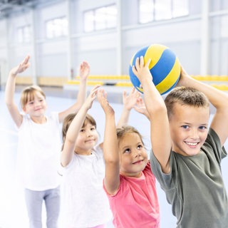 Kinder bei einem Ballspiel in einer Sporthalle: Zeitgemäßer Schulsport trainiert nicht nur verschiedene Sportarten, sondern auch soziale Fähigkeiten. Er motiviert Kinder und Jugendliche zur Bewegung.