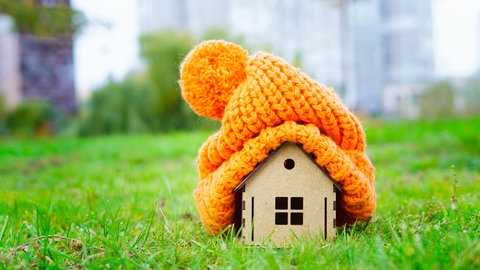 Holzhäuschen mit dicker orangefarbener Strickmütze auf dem Dach (Symbolbild) auf einer Wiese: Viele Wohnungen sind noch immer schlecht isoliert, Besitzer und Mieter heizen wortwörtlich zum Fenster hinaus; die energetische Sanierung kommt kaum voran. 