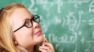 Mädchen mit Brille steht nachdenklich vor einer Kreidetafel mit Formeln: Intelligenz ist eine Art geistige Flexibilität, die sehr breit angelegt ist. Intelligenz ist eher allgemein, während Talente und Begabungen sich immer auf etwas beziehen. 