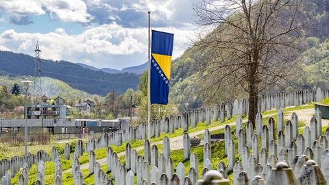 Eine bosnische Flagge weht auf Halbmast vor einer Reihe von Gräbern an der Gedenkstätte für den Völkermord von Srebrenica. Sie erinnert an die über 8.372 bosnischen Männer und Jungen, die von ihren Müttern, Schwestern und Töchtern getrennt und von bosnisch-serbischen Streitkräften Jugoslawiens getötet wurden.