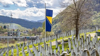 Eine bosnische Flagge weht auf Halbmast vor einer Reihe von Gräbern an der Gedenkstätte für den Völkermord von Srebrenica. Sie erinnert an die über 8.372 bosnischen Männer und Jungen, die von ihren Müttern, Schwestern und Töchtern getrennt und von bosnisch-serbischen Streitkräften Jugoslawiens getötet wurden.