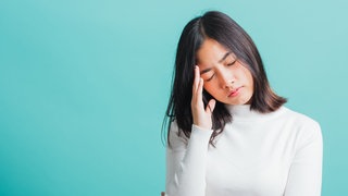 Junge Frau hat den Kopf zur Seite geneigt und hält sich die Schläfe: Ein Mensch leidet unter Schmerzen, Bluthochdruck oder Herzproblemen, aber die Ärztin findet keine körperlichen Ursachen, die das Ausmaß der Beschwerden erklären würden. Die Krankheit gilt dann als zumindest teilweise psychosomatisch bedingt.