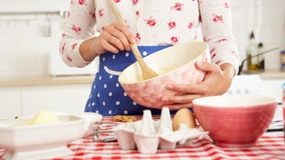 Junge Frau in Blümchenbluse und blauer Schürze rührt mit einem Holzlöffel in einer rosa Schüssel einen Kuchenteig an: Tradwife und Stayathomegirlfriend haben TikTok und Instagram erobert