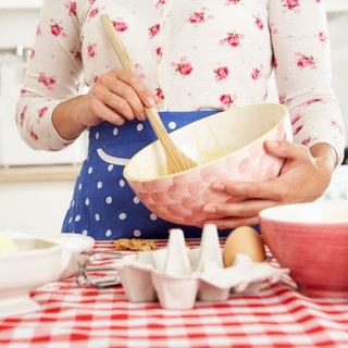 Junge Frau in Blümchenbluse und blauer Schürze rührt mit einem Holzlöffel in einer rosa Schüssel einen Kuchenteig an: Tradwife und Stayathomegirlfriend haben TikTok und Instagram erobert