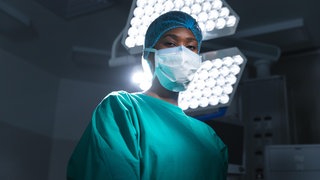 Schwarze Chirurgin mit Maske und OP-Kittel steht unter einer OP-Lampe: Krankenhäuser und Pflege-Einrichtungen sind auf medizinisches Personal aus dem Ausland angewiesen. 