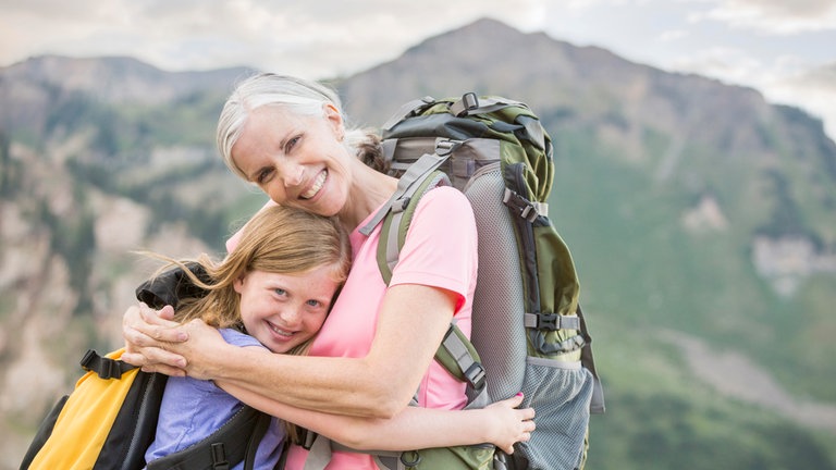 Großmutter und Enkelin mit Rucksäcken umarmen sich vor einem Bergpanorama: Wie wir schon als junge Menschen auf das Altwerden blicken, hat großen Einfluss darauf, wie wir selbst altern.