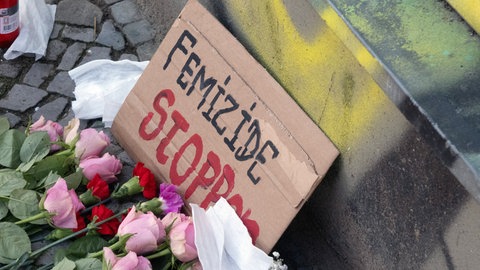 Blumen, niedergelegt für das Opfer eines Femizids. Auf einem Schild steht: "Femizide stoppen". Jedes Jahr versuchen in Deutschland geschätzt hunderte Männer ihre Partnerinnen und Ex-Partnerinnen zu ermorden, fast jeden zweiten Tag stirbt eine Frau.