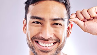 Junger Mann zupft lachend mit einer Pinzette eine Augenbraue: Wozu haben wir diese Haare über den Augen eigentlich?