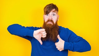 Junger Mann mit Bart zeigt "Daumen runter" und "Daumen rauf": Gutes Feedback kann Menschen sowohl im Beruf als auch im Privatleben weiterbringen. Doch wie gelingt Feedback und wie nimmt man Kritik an?