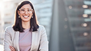 Junge Frau im Bewerbungsoutfit lacht mit verschränkten Armen in die Kamera: Viele Akademiker schreiben unzählige Bewerbungen – oft bleiben sie ohne Antwort. Wie gelingt der Berufseinstieg?