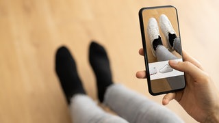 Eine Person probiert zu Hause auf dem Sofa mithilfe der App eines Versandhändlers virtuell neue Schuhe an: Wenn Kunden beim Online-Shopping mithilfe von KI passgenaue Mode auswählen können, soll das helfen, Retouren zu vermeiden.