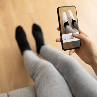 Eine Person probiert zu Hause auf dem Sofa mithilfe der App eines Versandhändlers virtuell neue Schuhe an: Wenn Kunden beim Online-Shopping mithilfe von KI passgenaue Mode auswählen können, soll das helfen, Retouren zu vermeiden.