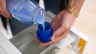 Jemand füllt blauen Weichspüler ins Weichspüler-Fach der Waschmaschine: Wie funktioniert der Weichspüler eigentlich?