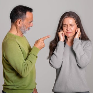 Wütender Mann zeigt mit Finger auf Frau; die hält sich die Ohren zu: Emotionale Gewalt ist oft schwer zu erkennen. Die Folgen sind jedoch gravierend. Gleichzeitig gilt: Nicht jedes böse Wort ist ein Übergriff. 