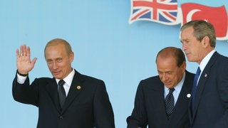 NATO-Russland-Rat: Russlands Präsident Wladimir Putin winkt im Beisein von US-Präsident George W. Bush (rechts) und Italiens Ministerpräsident Silvio Berlusconi nach Unterzeichnung eines Vertrages über eine neue Zusammenarbeit beim Russland-NATO-Gipfel am 28. Mai 2002 in Pratica di Mare bei Rom