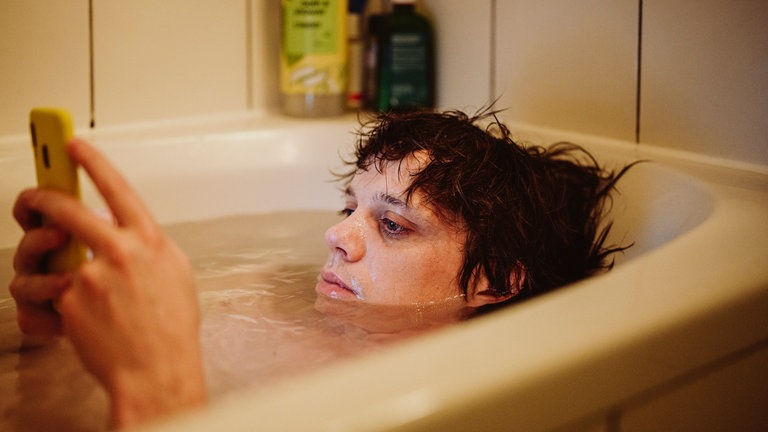 Zeno (Simon Steinhorst) in der Badewanne und scrollt durch sein Handy auf der Suche nach Kontakten.