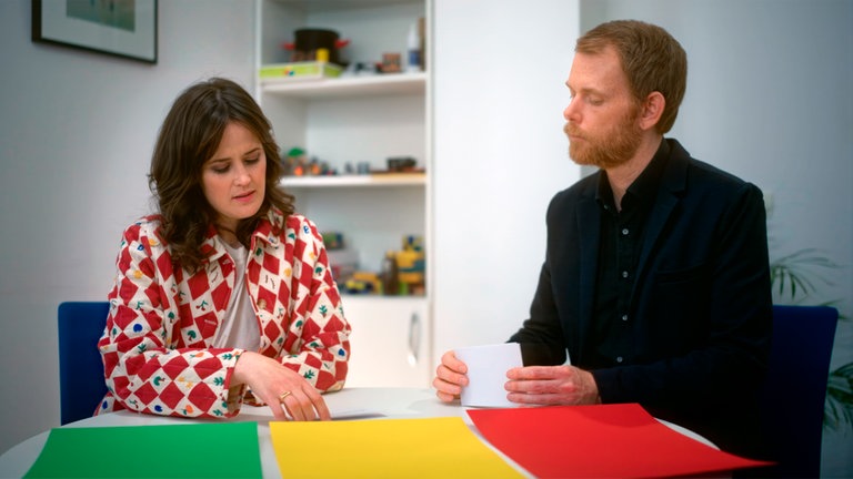 Julia Knörnschild sitzt mit dem Kinder- und Jugendpsychologen Prof. Julian Schmitz an einem Tisch. Vor ihnen liegen nebeneinander drei Bögen Papier in den Farben grün, gelb und rot. 