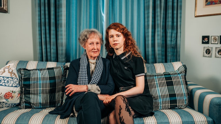 Luise (re.) und ihre Oma Guny sitzen nebeneinander auf der Couch und blicken ernst in die Kamera.