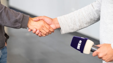 Zwei Personen reichen sich in einem hellen Innenraum die Hand: Links ist der Oberkörper einer Person in dunkelgrauem Pullover und Jeans zu sehen, rechts eine Person in einem hellgrauen, strukturierten Oberteil. Im Vordergrund unten rechts hält die rechte Person ein Mikrofon mit dunkelviolettem Windschutz, auf dem in weißer Schrift das Logo „SWR“ mit Pfeilsymbol steht, in Richtung des Handschlags.