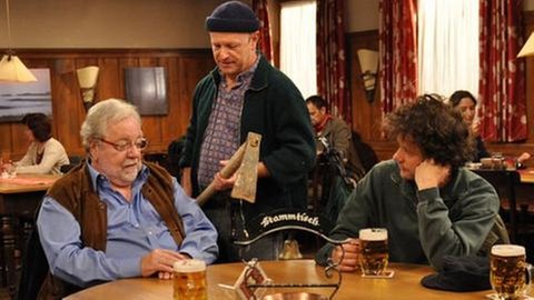 Hermann und Riedle sitzen am Stammtisch, Toni hält eine alte Hacke und steht neben ihnen