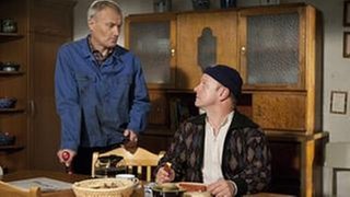 Toni sitzt in der Küche und isst, Karl steht auf eine Krücke gestützt neben ihm