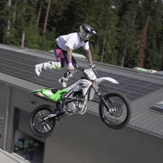 Freestyle-Motocrosser André Mikitta fliegt in waghalsiger Pose mit seinem Motorrad durch die Luft, vor blauem Himmel und unter Applaus des Publikums.