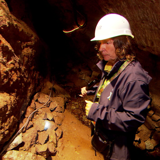 Die Bergwerksfrau steht mit einer Taschenlampe in einem dunklen Stollen der Grube Sophia und leuchtet die feuchten Felswände des historischen Bergwerks aus.