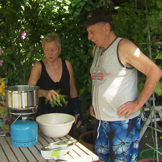 Ein Paar steht inmitten seines grünen Kleingartens, umgeben von Beeten und Sommerpflanzen, und freut sich über die Ernte.