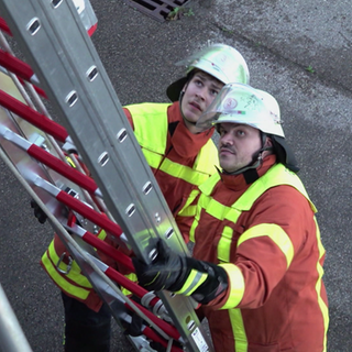 Zwei Feuerwehrmänner halten eine Leiter und schauen nach oben. Sie sind Teil einer Feuerwehrübung.