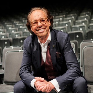 Kabarettist Christoph Sonntag sitzt in einem Raum voller Stühle in der ersten Reihe.