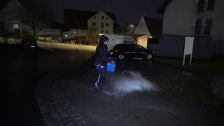 Auszubildender Amir ist mit einer schwarzen Jacke bekleidet. Er läuft nachts über eine Straße und streut Taumittel aus einem Eimer auf den Boden.