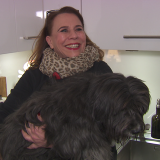 Eine Frau steht in einer Küche. Auf dem Arm hat sie einen Hund. Er ist fast schwarz und ein Tibet-Terrier-Dackelmix. 