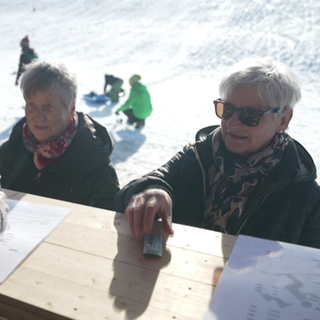 Zwei Frauen stehen an einer Après Ski-Hütte. Im Hintergrund Kinder die rodeln. 