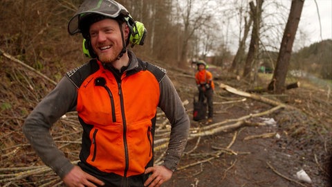 Philipp Klauser stet im Vordergrund und lacht, während er eine orangene Warnweste im Wald trägt.