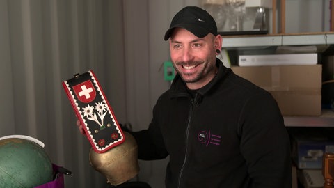 Nico lächelt in die Kamera und hat eine Kuhglocke mit der Schweizer Flagge in der Hand.