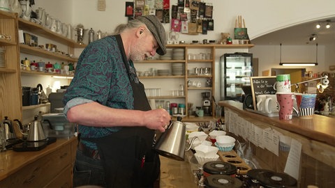 Ein Barista steht an einer hölzernen Theke und schüttet Kaffee aus einer metallenen Kanne in einen Kaffeefilter.