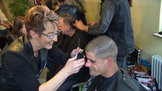 Eine Friseurin der Barber Angels lächelt und schneidet einem sitzenden Mann mit einem Rasierer die Haare.