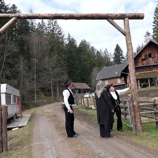Drei Menschen gehen durch einen hölzernen Torbogen. Links steht ein alter Wohnwagen. Rechts steht ein altes Bauernhaus.