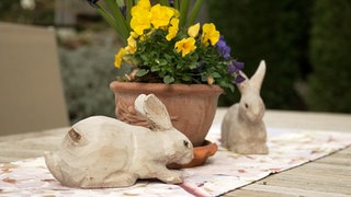 Zwei hölzerne Osterhasen sitzen auf einem Gartentisch. Zwischen ihnen ist ein Tontopf mit gelben und lianen Blumen.