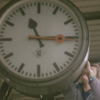 Eine große Uhr wird von einem Mann umgestellt. Er muss dafür auf eine Leiter steigen. 