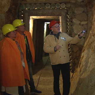 Eine Familie aus drei Personen steht in einem Bergwerk. Sie tragen orangene Umhänge und gelbe Helme. Ein Mann rechts im Bild leuchtet mit einer Taschenlampe.