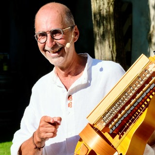 Bernhard Bitterwolf steht mit einem weißen Hemd draußen. In seiner Hand hält er eine hölzerne Drehleier. Er trägt eine Brille und ein Ohrenbügelmikrofon.