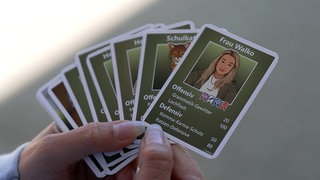 Eine Hand mit weißen Gelnägeln hält einige grüne Sammelkarten in der Hand. Auf der vordersten Karte steht der Name "Frau Walko", darunter eine Zeichnung von einer Frau mit blonden, langen Haaren.